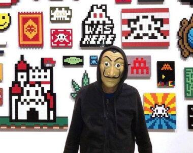 a photo of the french artist 'Invader', wearing his signature mask, stood in front of a wall filled with his artwork, Space Invader art