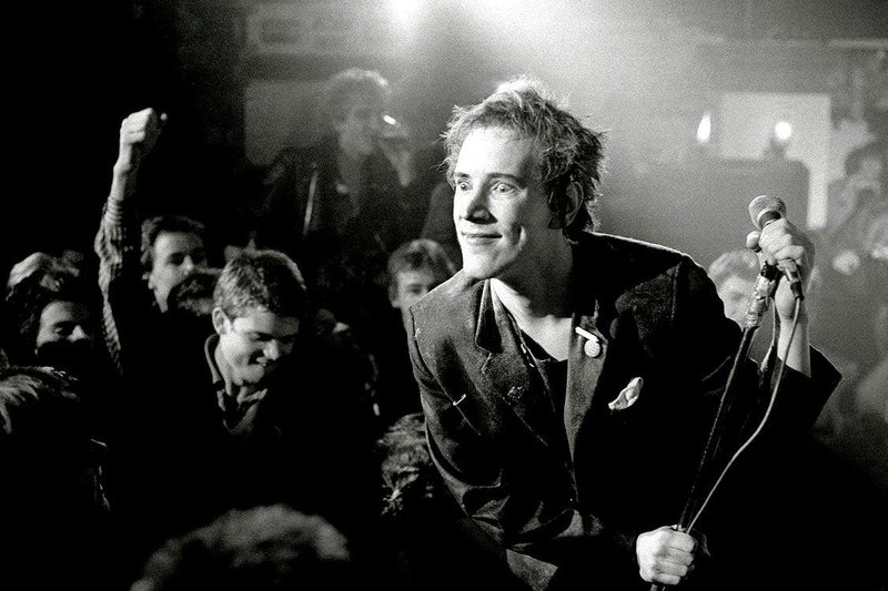 an image of a photograph by Kevin Cummins which is in black and white and features the singer of the sex pistols smiling into the crowd.