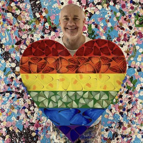 a photograph of artist damien hirst holding a piece of his artwork, the butterfly heart. the background features many colourful splodges of paint. the artwork is a heart shape in the colours of the rainbow featuring butterfly wings