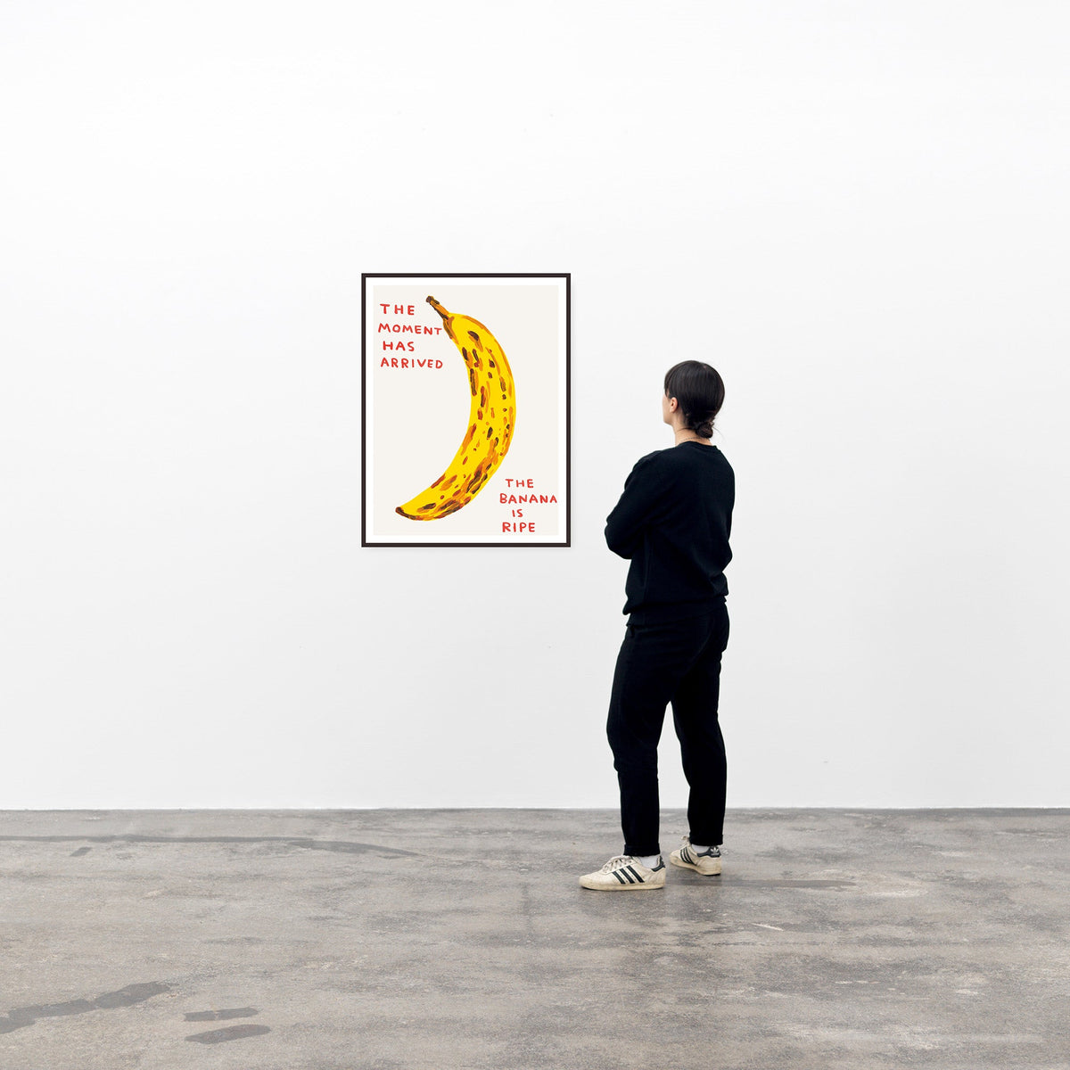 this is an image of a david shrigley artwork titled 'the moment has arrived' in a black frame, on a white wall with a viewer to its right. the artwork features a large painting of a brown spotted banana with handwritten text either side stating 'the moment has arrived', 'the banana is ripe'. this is a david shrigley print for sale