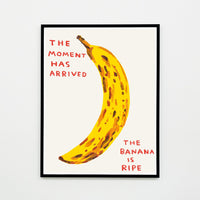 this is an image of a david shrigley artwork titled 'the moment has arrived' in a black frame. the artwork features a large painting of a brown spotted banana with handwritten text either side stating 'the moment has arrived', 'the banana is ripe'. this is a david shrigley print for sale