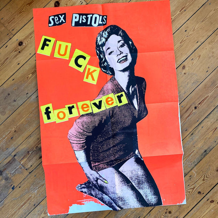 An image of the Jamie Reid artwork 'Fuck Forever', laid on a wooden floor. The artwork version has a red background, and lettering stating Sex Pistols Fuck Forever. 