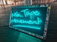 Experience the nostalgia of Mix Tape Movement by Tony Spink. This one-of-a-kind artwork features turquoise neon on a wooden backing panel painted black, adorned with a collage-style frame depicting mix tapes. Measuring 60 x 98 cm, this original masterpiece is a unique fusion of neon art and musical nostalgia. Elevate your space with this distinctive neon artwork capturing the essence of the mix tape era in a warehouse 