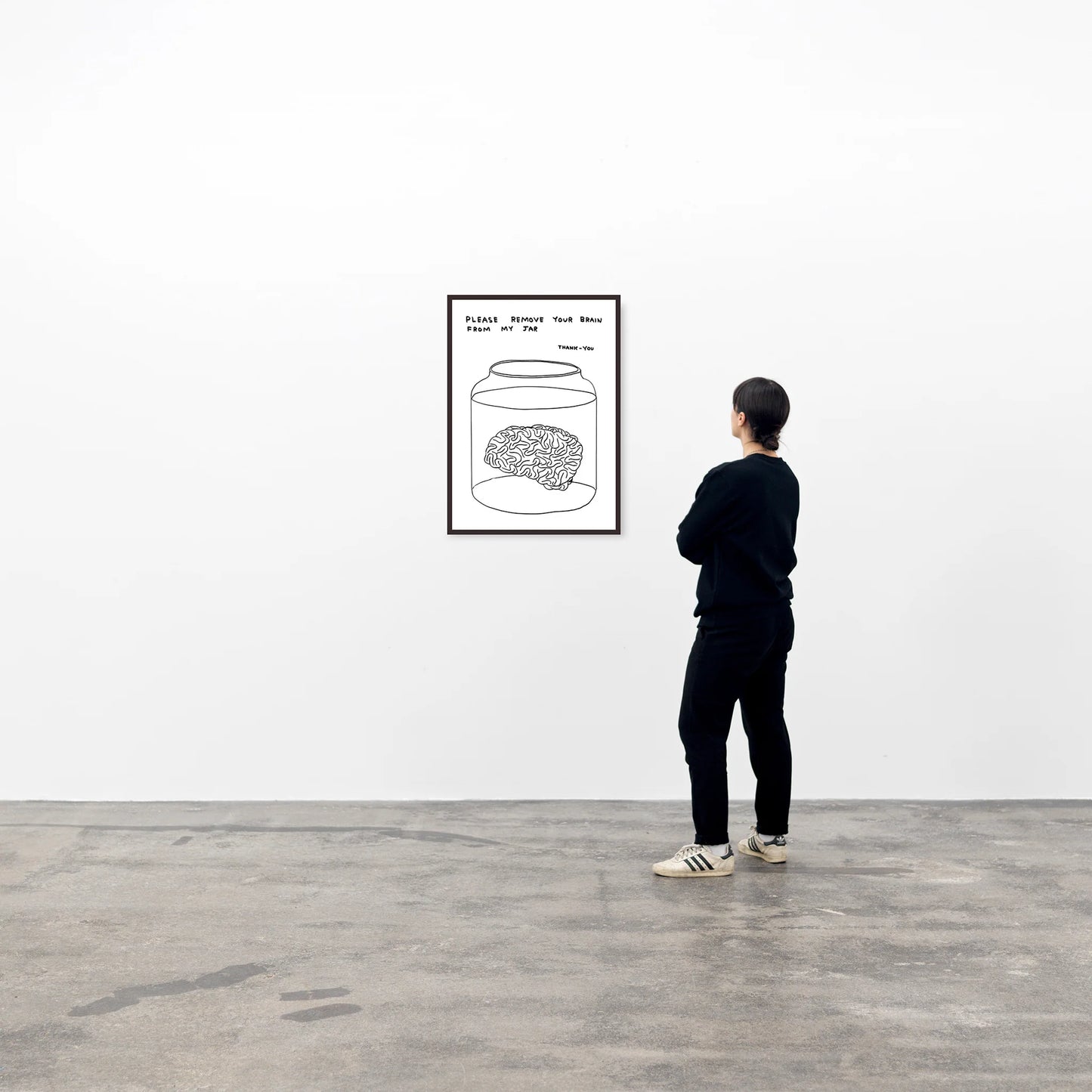 a photo of a david shrigley artwork titled 'please remove your brain from my jar' in a black frame, hung on a white wall and being looked upon by a single viewer on the right. the artwork features a white background, with a black line drawing of a brain in a jar of liquid, with text above stating 'please remove your brain from my jar tjank-you' in all caps. this is a david shrigley print for sale