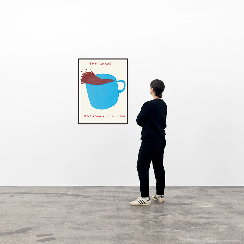 this is an image of a david shrigley artwork in a black frame on a wall with a single spectator to its right, and is untitled. the artwork features a blue teacup with coffee spilling out of it, with text stating 'THE CHAOS', 'EVENTUALLY IT WILL PASS'. this is a david shrigley poster for sale