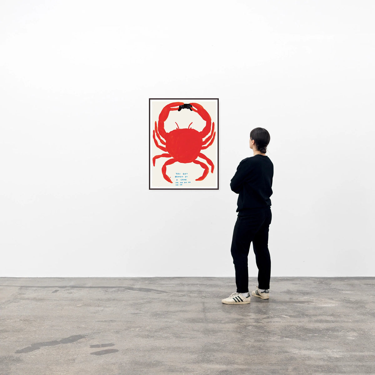 this is an image of an artwork by david shrigley titled 'you got beaten by a crab' in a black frame, hung on a white wall being viewed by a single spectator on its right. the artwork features a large orange crab holding a playstation controller, with blue text at the bottom stating 'YOU GOT BEATEN BY A CRAB HA HA HA HA HA HA'. this is a david shrigley poster for sale