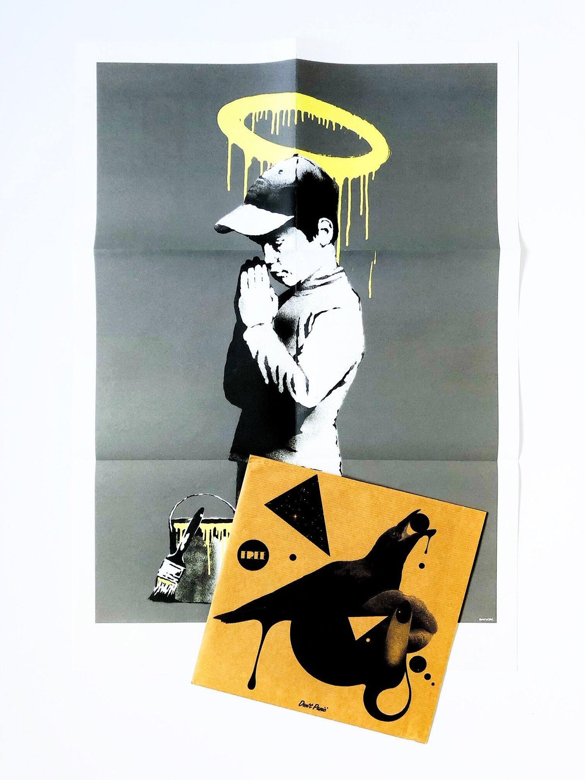 an image of an artwork by Banksy titled 'Forgive Us Our Trespassing', with a square image covering the bottom third of the artwork. The artwork features a stencilled image of a young boy in sportswear kneeling down and praying, with a yellow paintbucket in front of him, and a yellow halo painted on the wall above his head. this is a Banksy print for sale. 