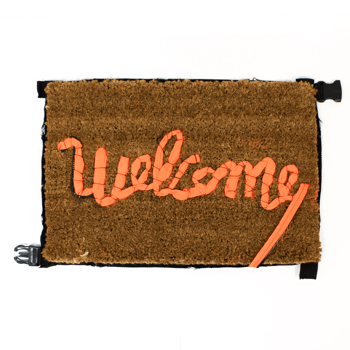 Buy Banksy merchandise online, Banksy for sale, Banksy Welcome Mat 2019, Banksy GDP 2019, Banksy Gross Domestic Product. Image is of a welcome mat, which is brown, stating 'welcome' in cursive writing. The item is distinctively rugged and rustic looking, with zips and safety belt buckles sticking out