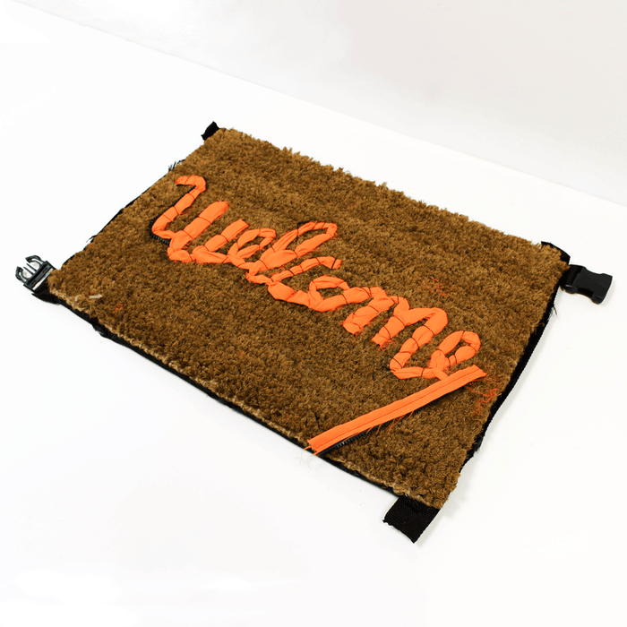 Buy Banksy merchandise online, Banksy for sale, Banksy Welcome Mat 2019, Banksy GDP 2019, Banksy Gross Domestic Product. Image is of a welcome mat, which is brown, stating 'welcome' in cursive writing. The item is distinctively rugged and rustic looking, with zips and safety belt buckles sticking out